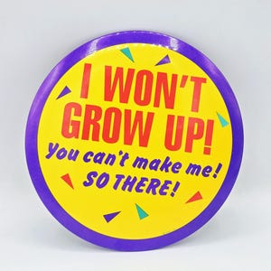 May include: A circular badge with a yellow centre and a purple border. The badge displays the text "I WON'T GROW UP! You can't make me! SO THERE!" in red and blue. Small triangles in green, blue, and red decorate the badge.