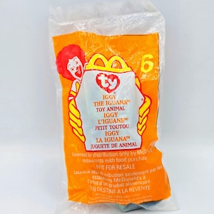 May include: A yellow and red Ty Beanie Baby toy packaging featuring a picture of Ronald McDonald. The packaging states "IGGY THE IGUANA" and "TOY ANIMAL".