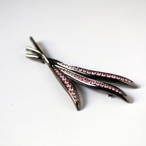 May include: A silver-toned brooch in a stylized leaf design. The brooch features three curved elements, each lined with small, pink gemstones. The pin is set against a white background, highlighting the contrast of colors and textures.