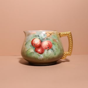 May include: A ceramic jug with a hand-painted design of red apples and green leaves. The handle is gold and decorated with a beaded pattern. The jug is a decorative item. The jug is a decorative item.