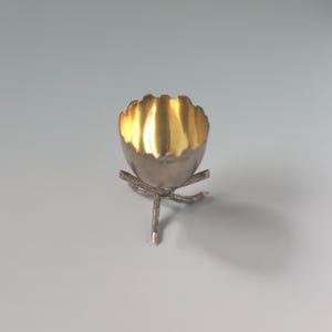 Pode incluir: Um porta-ovos prateado com interior dourado. O porta-ovos tem uma borda ondulada e assenta num suporte de três pernas. O suporte tem um design semelhante a um galho.