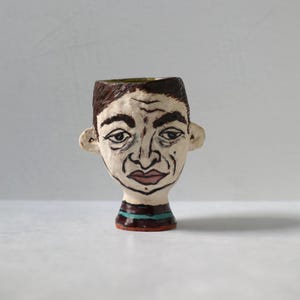 May include: A handmade ceramic vase shaped like a human head. The face is painted with dark brown hair, black eyebrows, and a red-brown mouth. The neck has a striped pattern of brown and teal. The vase is a unique piece of art.
