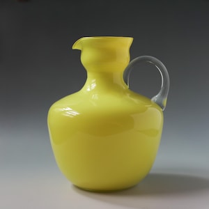 Op de afbeelding: Een heldergele glazen kan met een transparante glazen handvat. De kan heeft een rond lichaam en een licht uitlopende lip.