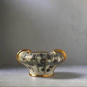 May include: A small, decorative ceramic bowl with a cream-colored base and gold trim. The bowl features a floral design in shades of blue and green. The handles are also trimmed in gold.