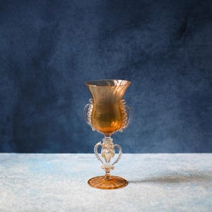 May include: An amber-colored glass goblet with a flared rim and two ornate handles. The goblet features a detailed, clear glass stem and a circular base. The background is a textured blue surface.