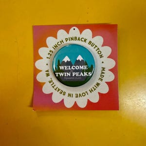 May include: A 1.25-inch pinback button with a teal and white mountain scene and the text "WELCOME to TWIN PEAKS" on a white daisy-shaped card. The card also reads "MADE WITH LOVE IN SEATTLE, WA."