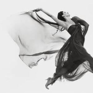Könnte beinhalten: Eine Frau in einem schwarzen Kleid mit wallendem Rock springt in die Luft. Das Kleid ist aus einem durchsichtigen Stoff, der im Wind weht. Die Frau trägt hohe Absätze und hat ihre Arme ausgestreckt.