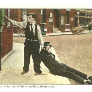 Puede incluir: Una fotografía vintage en blanco y negro de Buster Keaton en una de sus comedias. Keaton está vestido con traje y corbata, y está sujetando a un hombre con traje y gorra. El hombre está tumbado en el suelo, y Keaton está mirando a la cámara. La foto está ambientada en una calle de la ciudad, con edificios en el fondo.