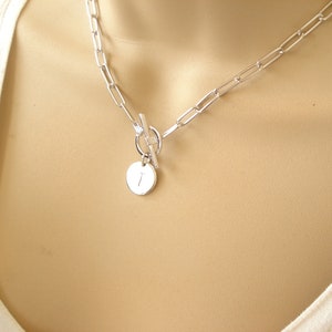 May include: A silver chain necklace with a toggle clasp and a round pendant with the letter "I" engraved on it.