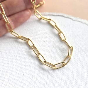 May include: A gold-colored chain necklace with elongated rectangular links. The chain is displayed on a white surface, with a hand partially visible in the background.