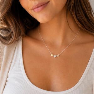 Puede incluir: Un collar de cadena plateada fina con tres pequeños dijes en forma de corazón dispuestos horizontalmente. Los dos corazones exteriores son de tono dorado, mientras que el corazón central es de tono plateado. El collar descansa sobre la clavícula.