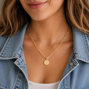 Op de afbeelding: Een goudkleurige ketting met een ronde hanger waarin de letter J is gegraveerd. De hanger hangt aan een ketting die bestaat uit langwerpige, ovale schakels. De ketting rust tegen de hals en toont de metalen afwerking en het ontwerp.