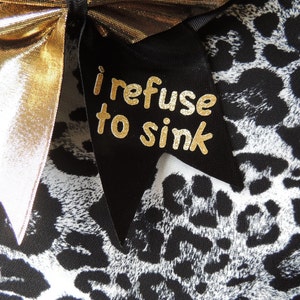 May include: A black and white leopard print fabric with a gold bow and a black ribbon with the text "i refuse to sink" in gold letters.