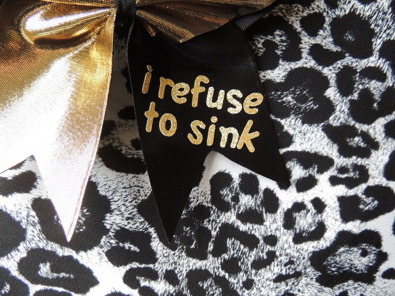 May include: A black and white leopard print fabric with a gold bow and a black ribbon with the text "i refuse to sink" in gold letters.