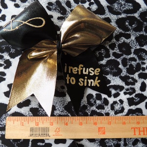 May include: A black and gold cheer bow with a white center. The bow has the words "I refuse to sink" written on it in gold glitter. The bow is on a black and white leopard print fabric.