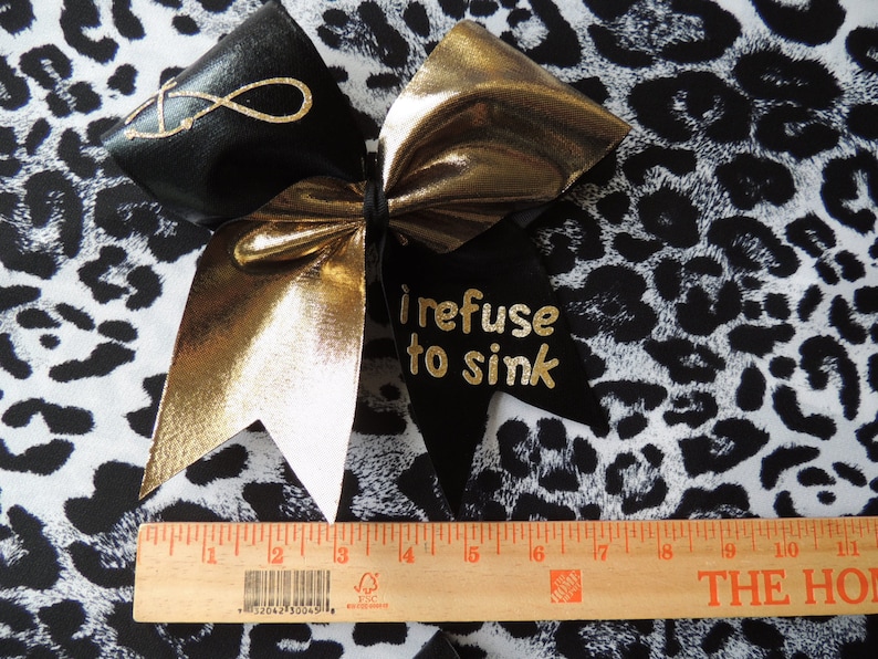 May include: A black and gold cheer bow with a white center. The bow has the words "I refuse to sink" written on it in gold glitter. The bow is on a black and white leopard print fabric.