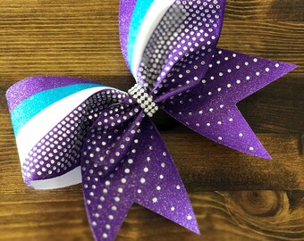 May include: A purple and white cheer bow with teal stripes and silver glitter. The bow is decorated with rhinestones.