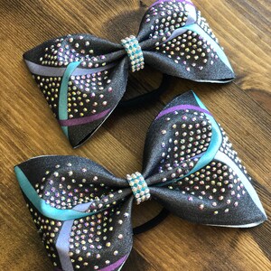 May include: Two black and grey cheer bows with a teal, purple and grey geometric pattern. The bows are decorated with iridescent rhinestones and have a black elastic band.