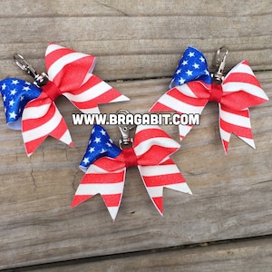 May include: Three red, white and blue glitter bow clips with silver swivel clips. The bows are decorated with a glittery American flag pattern. The website www.bragabit.com is printed on the wooden surface.