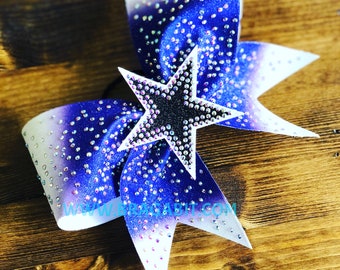 Puede incluir: Un lazo de animadora blanco y azul degradado con una estrella negra y plateada con purpurina en el centro. El lazo está decorado con pedrería iridiscente.
