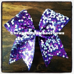 May include: Purple and white cheer bow with sequins. The bow is tied in a large, double loop with a center knot. The bow is decorated with a variety of purple and white sequins. The text "@braggaboutcheerbows" is printed on the bow.