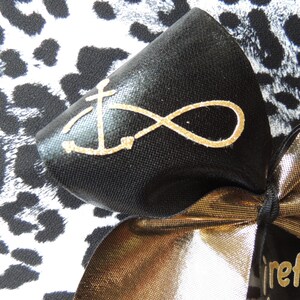 May include: A black bow with a gold infinity symbol and anchor design on a black and white leopard print background. The bow is tied with a gold ribbon and has a tag that says "refuse to sink".
