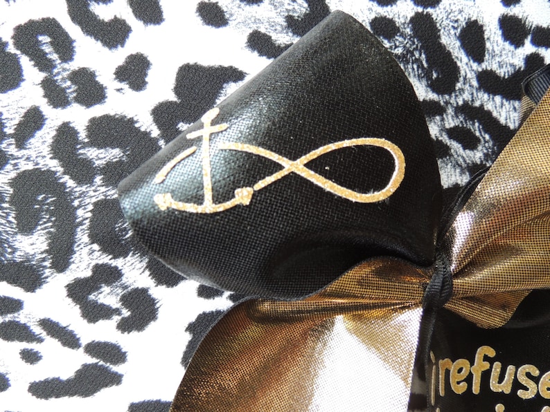 May include: A black bow with a gold infinity symbol and anchor design on a black and white leopard print background. The bow is tied with a gold ribbon and has a tag that says "refuse to sink".