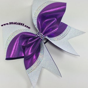 Op de afbeelding: Paarse en witte cheer bow met strass. De strik is gemaakt van glanzend materiaal en heeft een witte glitterafwerking op de uiteinden. De strik is vastgebonden met een zwart elastiek.