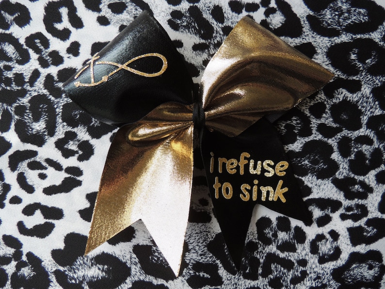 May include: A black and gold cheer bow with the text "I refuse to sink" on one side and an infinity symbol on the other side.