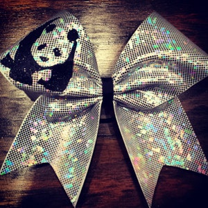 Può includere: Un fiocco da cheerleader bianco e argento con un design di panda nero. Il fiocco è realizzato in materiale iridescente con una finitura scintillante.