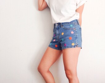 May include: A pair of denim shorts with colorful embroidered flowers. The shorts are a light blue wash and have a high waist.