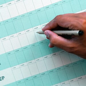 May include: A close-up of a teal and white monthly calendar with the months of May through September visible. A hand holds a pen, poised to write on the calendar. The calendar is used for planning and scheduling.
