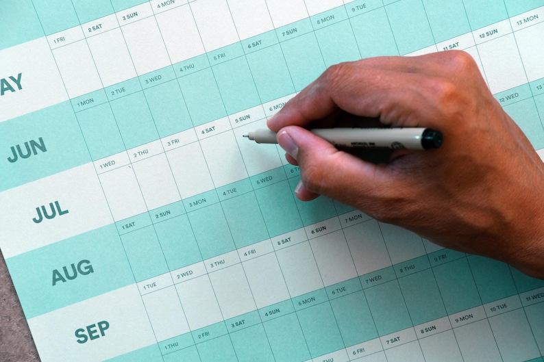May include: A close-up of a teal and white monthly calendar with the months of May through September visible. A hand holds a pen, poised to write on the calendar. The calendar is used for planning and scheduling.