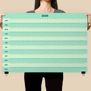 May include: A large, rectangular 2026 year calendar in shades of green and white. The calendar is held up by a person. The months are listed vertically on the left side. The calendar is secured with black binder clips.