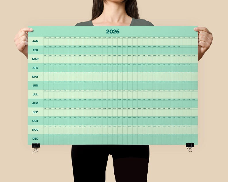 May include: A large, rectangular 2026 year calendar in shades of green and white. The calendar is held up by a person. The months are listed vertically on the left side. The calendar is secured with black binder clips.