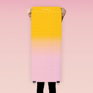 May include: A long, rectangular 2026 calendar with a gradient design, transitioning from yellow to pink. The calendar is held up by a person, with only their hands and legs visible. The calendar is secured with binder clips.