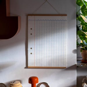 May include: A white wall calendar with a wooden frame and a string hanging from the top. The calendar is blank and has a grid pattern. The calendar is hanging on a white wall with a brown shelf and a plant in a pot on the right side of the image.