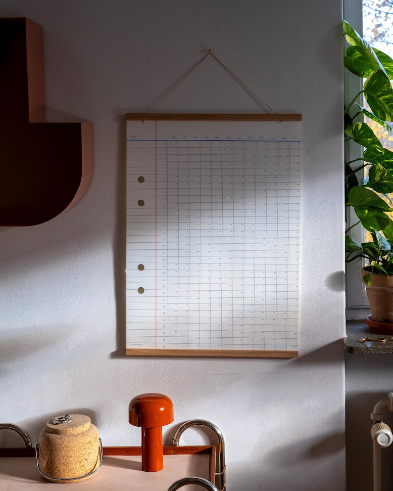 May include: A white wall calendar with a wooden frame and a string hanging from the top. The calendar is blank and has a grid pattern. The calendar is hanging on a white wall with a brown shelf and a plant in a pot on the right side of the image.