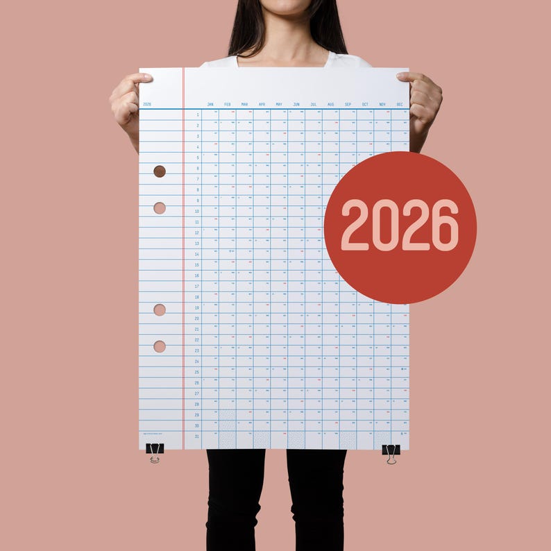 May include: A person is holding up a large white sheet of paper with a grid pattern and the year 2026 written in white on a red circle. The paper has three small brown dots on the left side.