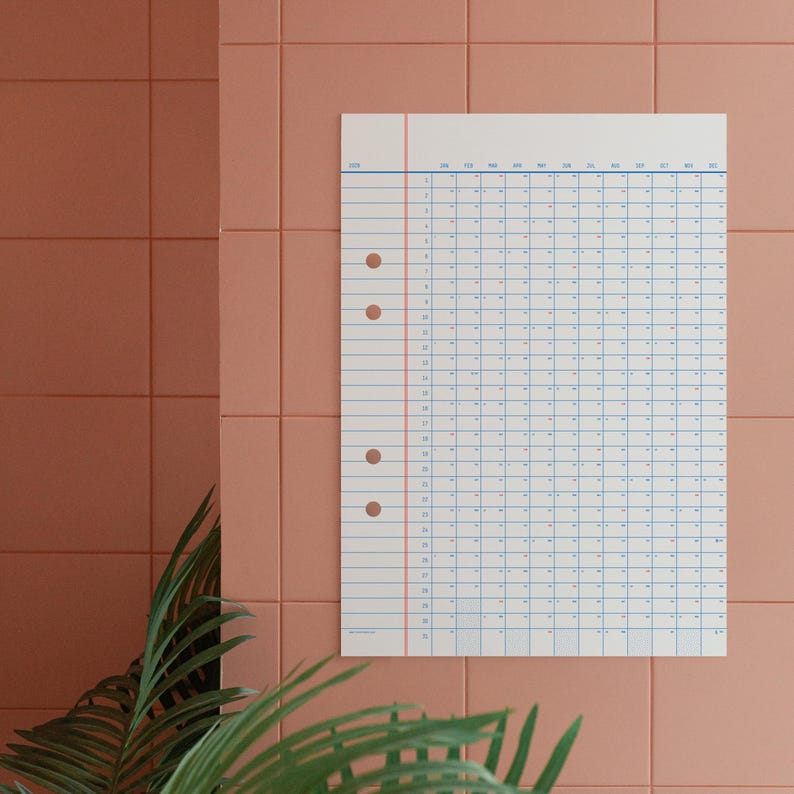 May include: A white calendar with blue and red lines, hanging on a pink tiled wall. The calendar is a grid with numbers 1 through 31 for each month of the year. The calendar is labelled with the year 2023.