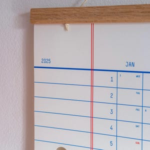 May include: A white wall calendar with a wooden frame and red and blue lines. The calendar is for the year 2025 and the month of January. The days of the week are labelled in blue and the numbers for the days of the month are in black.