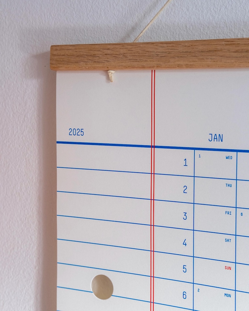 May include: A white wall calendar with a wooden frame and red and blue lines. The calendar is for the year 2025 and the month of January. The days of the week are labelled in blue and the numbers for the days of the month are in black.