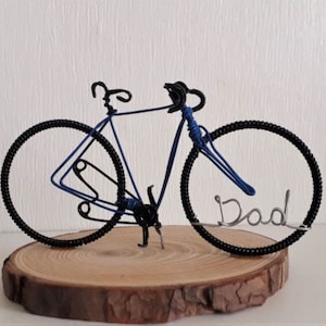 Wire sculptured bicycle. In the style of a racer. It is painted with a choice of nine colours, personalised with a name written in wire and set into the front wheel. The bicycle is approximately nine to ten cm long and is set onto a wood cutting