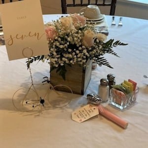 Può includere: Un cartoncino bianco per il numero del tavolo con la parola "sette" scritta in calligrafia dorata. Il cartoncino è tenuto da un supporto per bicicletta in filo metallico. Una scatola di legno piena di fiori rosa e bianchi è posta sul tavolo. I fiori sono circondati da verde.