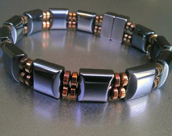 May include: A black hematite bracelet with copper and gold accents. The bracelet is made of square-shaped beads.