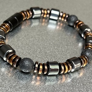 Puede incluir: Un brazalete de cuentas negras y cobre con cuentas de lava negra. El brazalete está hecho con cuentas redondas y cilíndricas.