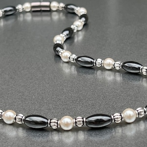 May include: A black and white beaded necklace with a magnetic clasp. The necklace is made with oval black beads, white pearls, and silver spacer beads.