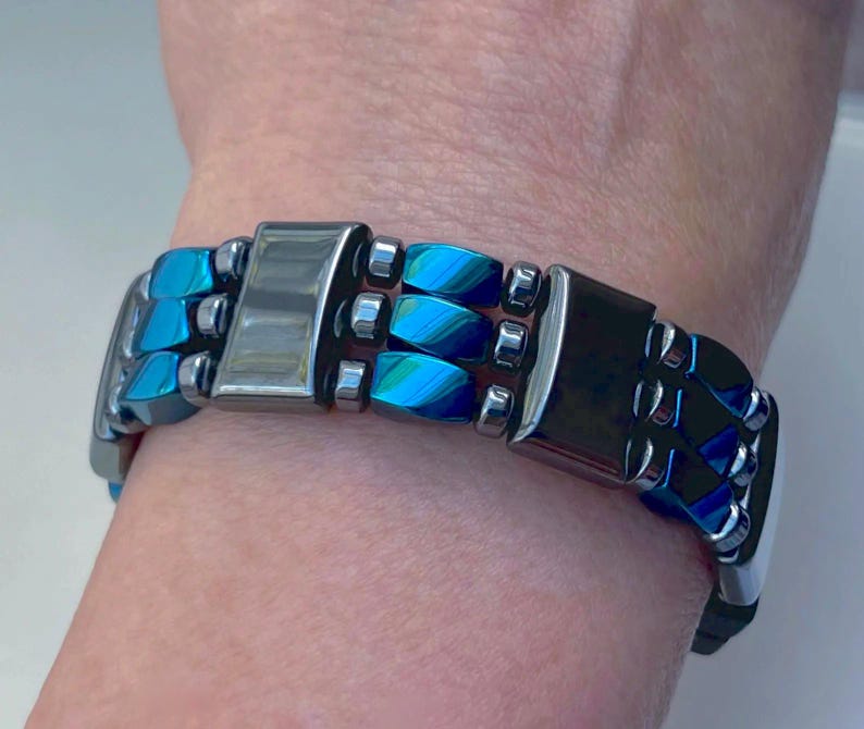 May include: A blue and silver magnetic bracelet with a clasp closure. The bracelet is made of multiple rows of beads.