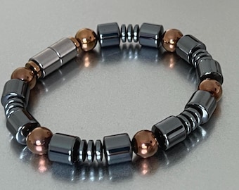 May include: A bracelet featuring a mix of dark gray, cylindrical, and square beads, interspersed with copper-colored spherical beads. The bracelet has a magnetic clasp and is designed to be worn on the wrist.