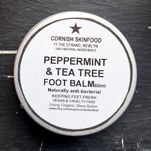 May include: A silver tin with a white label that reads "Cornish Skinfood" and "Peppermint & Tea Tree Foot Balm 60ml". The label also states that the product is "Naturally anti-bacterial", "Keeping Feet Fresh", "Vegan & Cruelty Free", and "Using Organic Shea Butter".